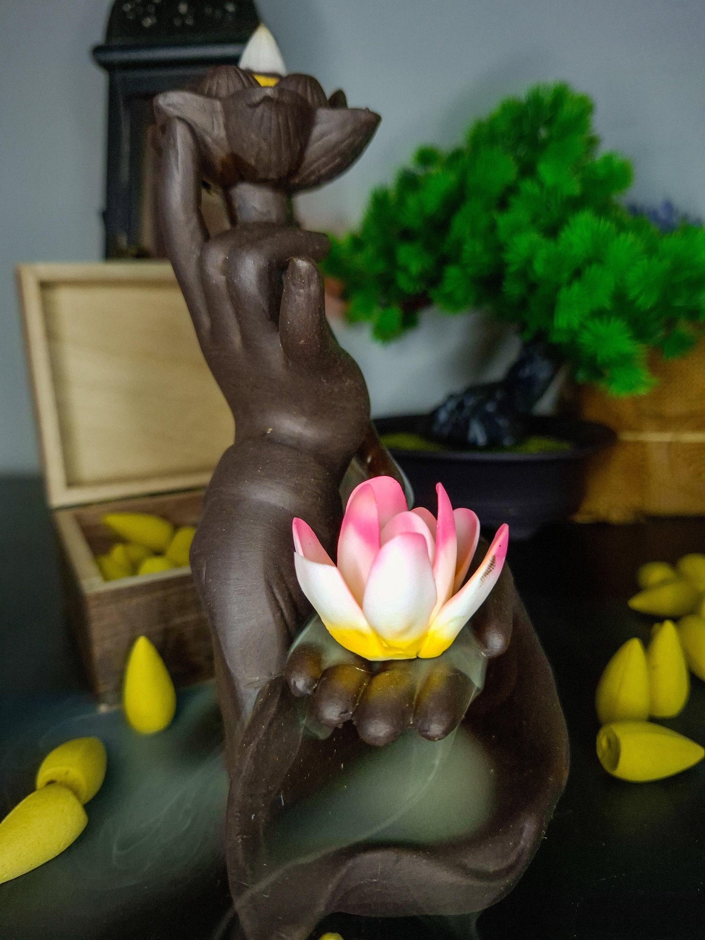Incense burner shaped like a hand holding a pink lotus flower, with incense cones and smoke.